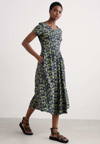 Rochie florală bleumarin cu mâneci scurte, având un corset ajustat, fustă evazată, buzunare și modele florale galben-verzi. Asortată cu sandale negre.