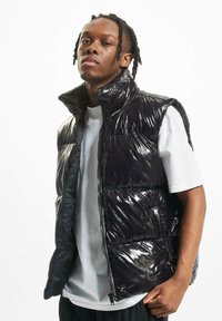 Black shiny puffer vest with a high collar, zip closure, and structured sections. Worn over a white T-shirt, showcasing a glossy texture.