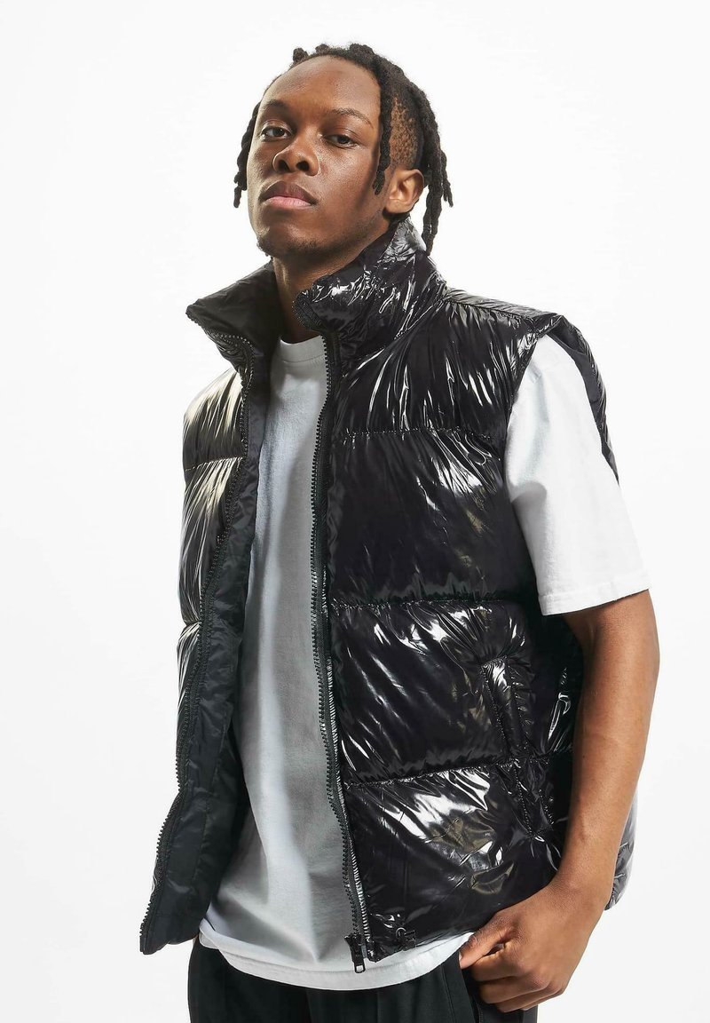 Black shiny puffer vest with a high collar, zip closure, and structured sections. Worn over a white T-shirt, showcasing a glossy texture.