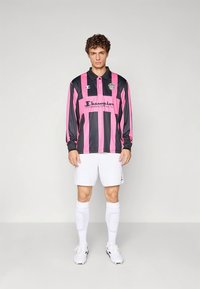 En ung man står iklädd en långärmad tröja med rosa och svarta ränder och "Champion"-logotyp, vita shorts, strumpor och fotbollsskor mot en enkel bakgrund.