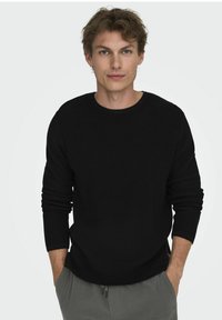 Schwarzer Strickpullover mit Rundhalsausschnitt und langen Ärmeln. Model trägt graue strukturierte Hose, mit Händen in den Taschen, vor einem neutralen Hintergrund.