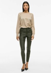 Beige satinvask langermet bluse med rynkede mansjetter, kombinert med olivengrønne smale bukser; modellen har på seg svarte spisse hæler.