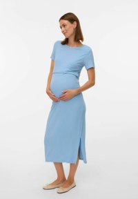 Robe de maternité bleu clair à rayures, avec des manches courtes, une fente sur le côté, et une coupe ajustée qui met en valeur un ventre rond. Des chaussures plates simples complètent le look.