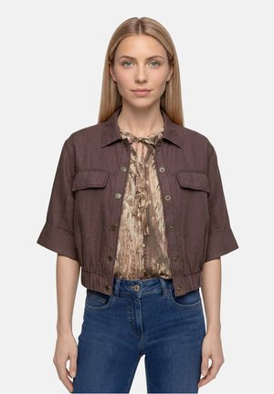 Femme aux cheveux blonds et raides porte une veste courte marron par-dessus un chemisier à motifs et un jean bleu, debout devant un fond uni.
