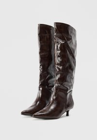 Botas oscuras de cuero con patrón de cocodrilo, hasta la rodilla, con punta afilada y tacones bajos tipo kitten sobre un fondo blanco sencillo.