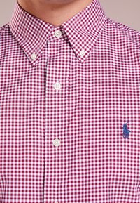 Czerwona i biała koszula w kratkę gingham z kołnierzykiem zapinanym na guziki, białymi guzikami i niebieskim haftowanym logo na lewej piersi.