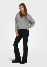 Pull en maille gris avec un col en V, porté par-dessus un haut en dentelle noir, associé à un pantalon évasé noir et des bottes noires à semelles épaisses.