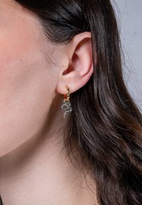 Guldörhänge med en blå, ormsformad charm med texturerad detaljering, hängande från ett öra med mörkt hår synligt.