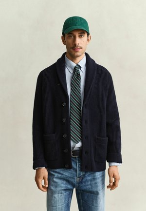 Mann trägt grüne Kappe, marineblauen Cardigan, hellblaues Hemd mit Knöpfen, grün gestreifte Krawatte und blaue Jeans, steht vor einfachem Hintergrund.