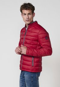 Rød pufferjakke med høj krave, kviltet tekstur, frontlynlås og kontrastfarvede marineblå detaljer. Slim fit med et rundt emblem på ærmet.