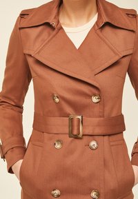 Hnědý dvouřadý trench coat s opaskem v pase, velkým límcem a knoflíky ve zlaté barvě. Texturovaná látka s rozloženým designem.