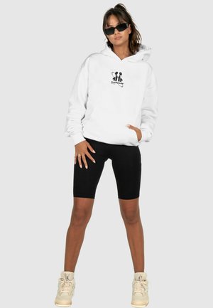 Weißer Hoodie mit einem schwarzen Grafikdesign, kombiniert mit schwarzen Radshorts und beigen Sneakers. Das Modell trägt schwarze Sonnenbrillen.