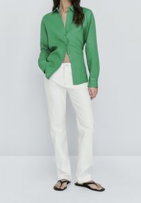 Woman wearing a green buttoned shirt, white straight-leg pants, and black flip-flops, standing against a plain light background.