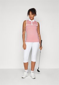 Polo skjorte i lys rosa med hvite detaljer, ermeløs design, krage med knapper. Hvit capribukse og golfkølle i hånden.