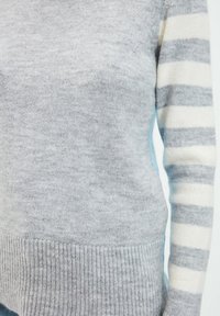 Maglione grigio a maglia con orlo a coste, una manica presenta strisce bianche e grigie. Il materiale sembra morbido, con una trama confortevole.