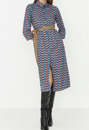 Kobieta ubrana w wzorzystą sukienkę midi z żółtym paskiem we wzory przewiązanym w talii, do tego nosi czarne kozaki za kolano.