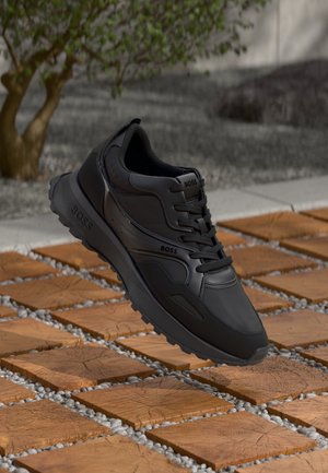 Baskets noires de la marque Black Boss avec lacets sur des dalles carrées en bois entourées de petits cailloux en extérieur près d’un arbre.