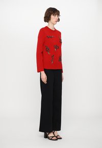 Red knitted sweater with snake patterns, ribbed hem, paired with black wide-leg trousers and black heeled sandals. Side view composition.