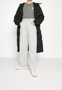 Schwarze Puffjacke, graues T-Shirt mit Grafik, hellblaue weit geschnittene Jeans und weiße Sneaker mit Gummisohlen. Einfaches und lässiges Outfit.