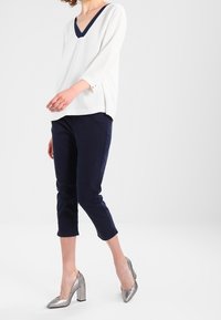 Personne portant un blouse blanche en V, un pantalon cropped navy et des chaussures à talons argentées, se tenant avec un pied légèrement en avant.