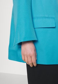 Tyrkysový blejzr s hladkou texturou, který má široký rukáv a jednu kapsu blízko spodního lemu, kombinovaný s černým oděvem pod ním.