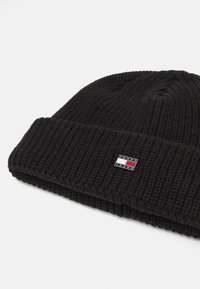 Gorro de punto negro con textura acanalada y un dobladillo doblado. Presenta una pequeña etiqueta de marca en rojo, blanco y azul en la parte frontal.