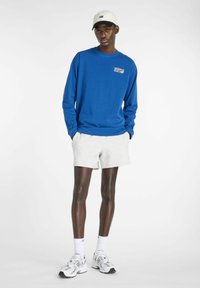 Camicia a maniche lunghe blu con logo stampato, abbinata a pantaloni corti grigi. Il modello indossa un cappellino bianco e sneakers New Balance, mostrando un outfit casual.