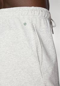 Šviesiai pilki medvilniniai sportiniai džinsai su elastine juosta ir raišteliu. Yra šoninės kišenės ir mažas žalias logotipas priekyje.