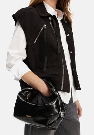 Donna che indossa un giubbotto senza maniche nero sopra una camicia bianca, che tiene in mano una borsa nera lucida con logo dorato.