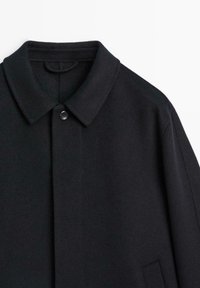 Cappotto in lana nera con collo a revers, chiusura con bottoni e tasca angolata. Presenta una superficie liscia e un design strutturato.