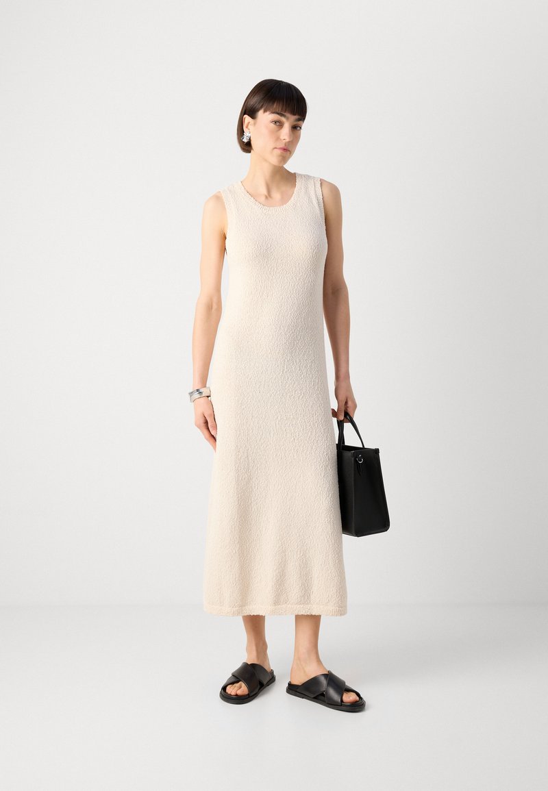 Robe tricot sans manches beige avec une finition texturée, sac à main noir et sandales à glisser noires. Conception simple, longueur mi-mollet.