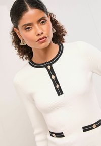 Fehér bordázott kötött felső fekete részletekkel, kerek nyakkivágással, három arany gombbal és zsebekkel, melyeket fekete csíkozás díszít.