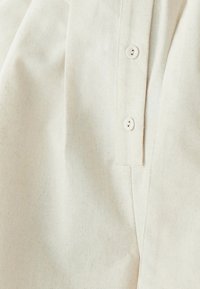Béžová bavlněná košile s knoflíkovou légou, jemnou texturou a měkkým materiálem. Design zahrnuje sklopený límec a rovné švy.