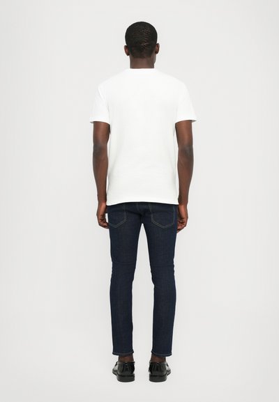 Una camiseta blanca de manga corta combinada con unos jeans oscuros de corte ajustado y zapatos negros, vista desde la parte de atrás contra un fondo neutro.