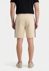 Hombre con pantalones cortos beige con franjas negras laterales, camiseta negra y zapatillas beige, de pie de espaldas contra un fondo liso.