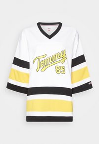 Bílý dres s žlutým nápisem "Tommy" a číslem "85". Rukávy a tělo zdobí černé a žluté pruhy. V-výstřih.