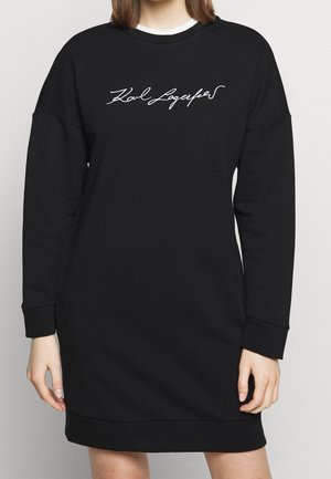 Kvinde iført en sort langærmet kjole med "Karl Lagerfeld" skrevet med hvid skrift på brystet, stående foran en ensfarvet baggrund.