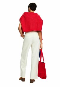 Châle en tricot rouge drapé sur les épaules, porté avec un pantalon blanc à jambes larges et des chaussures marron à talons blocs. Tenant un sac fourre-tout rouge.