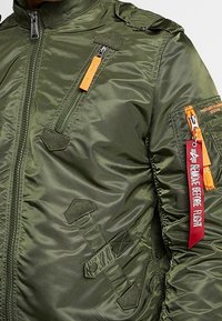 Blouson aviateur vert olive à finition brillante, doté de plusieurs poches zippées, d'un écusson, et d'une étiquette orange "Remove Before Flight".