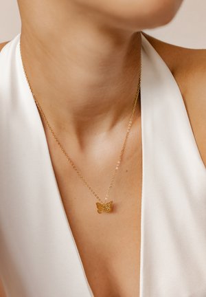 Goldener Schmetterling-Anhänger mit feiner Kette, vor einem hellen Oberteil präsentiert. Mit filigranen Details auf dem Schmetterling.