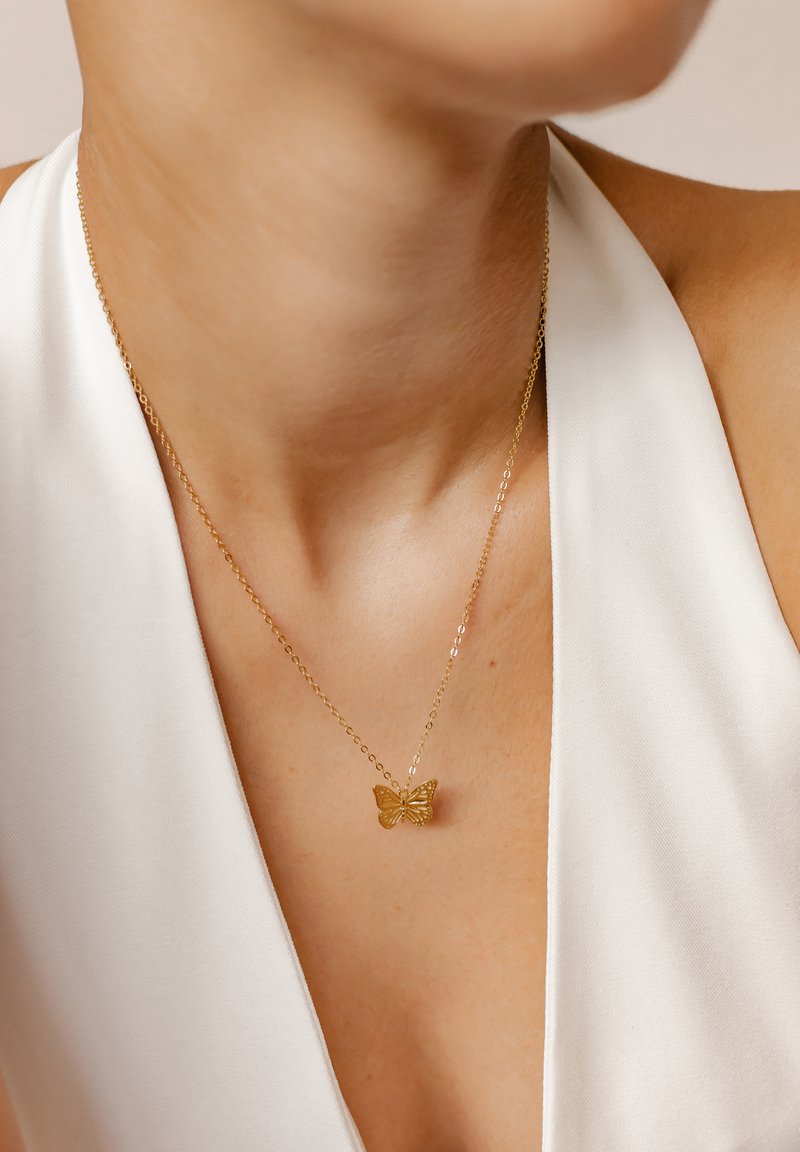 Collar con colgante de mariposa dorada y una cadena delicada, sobre una blusa de color claro. Presenta un detallado intrincado en la mariposa.