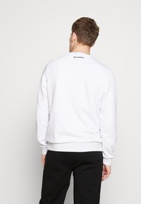 Sudadera blanca con cuello redondo y puños acanalados, que presenta un logo negro de "KARL LAGERFELD" en la parte posterior. Textura de tela suave.