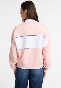 Sudadera rosa con una franja horizontal blanca y acentos azules. El diseño tiene un corte holgado y puños acanalados, combinada con jeans azules.