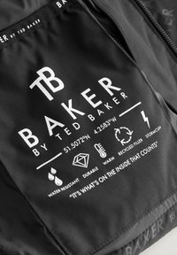 Crna tkanina oznaka s bijelim tekstom koji sadrži informacije o vodootpornosti, izdržljivosti, toplini, recikliranom punilu i manžetama otpornih na oluju. Logo "Baker by Ted Baker."