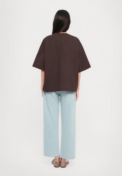 T-shirt oversize marrone, tessuto leggero, maniche corte, abbinato a pantaloni a gamba larga in denim chiaro con orlo sfilacciato. Le calzature presentano strisce multicolori.