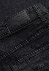 Jean en denim noir avec une étiquette en cuir portant le logo de la marque « ombre ». Le tissu présente une finition texturée avec des détails de couture visibles.
