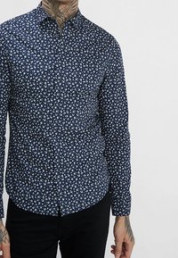 Camicia blu navy a maniche lunghe con bottoni, motivo floreale bianco, dal design aderente e colletto semplice. Tessuto texture.