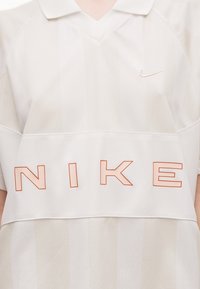 Λευκό πόλο μπλουζάκι Nike με διακριτικές κάθετες ρίγες, μεγάλο πορτοκαλί περίγραμμα του κειμένου «NIKE» στο στήθος και μικρό λογότυπο swoosh στο πάνω δεξιά μέρος.