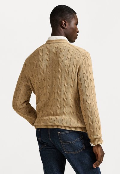 Gaiši brūns adīts džemperis ar ribotiem piedurknēm un apakšu, ar rakstainiem tekstūras elementiem. Saskaņots ar tumši ziliem džinsa biksēm.