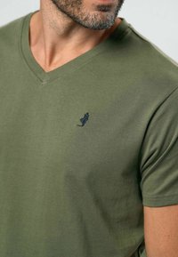 Alyvuogių žalios spalvos medvilniniai marškinėliai su V formos iškirpte ir trumpomis rankovėmis. Kairėje krūtinės pusėje yra mažas tamsiai mėlynas siuvinėtas logotipas.